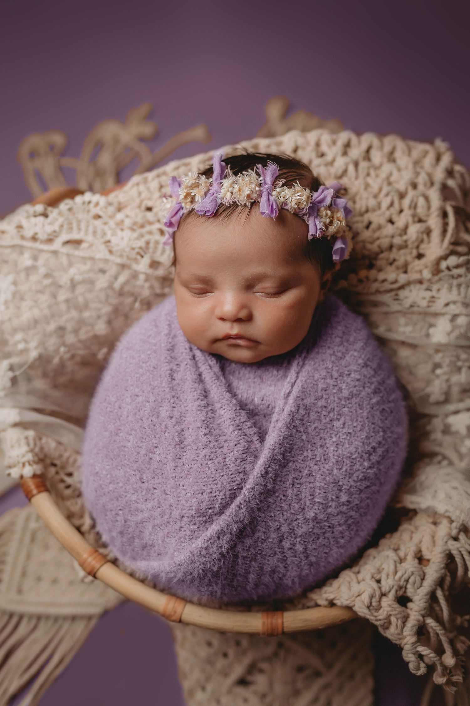 Sleeping newborn wrapped in purple swaddle wearing delicate floral crown.