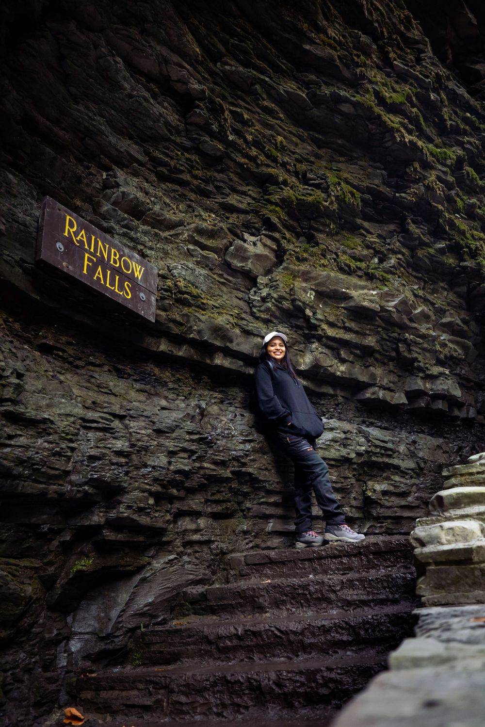 Adventure seeker poses beneath Rainbow Falls sign on dramatic rocky cliff face.
