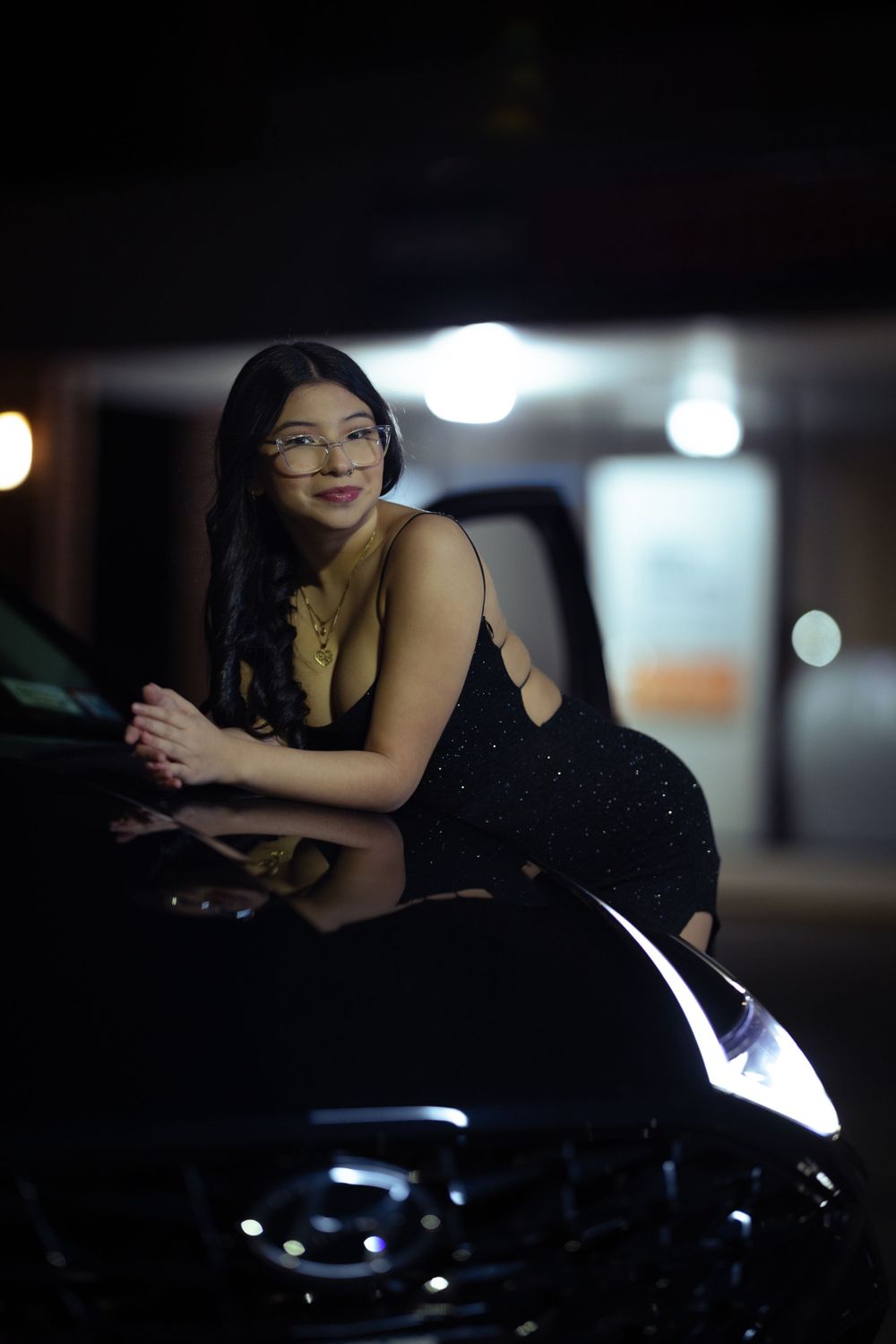 Woman in sparkly dress poses on black car under nighttime lighting.