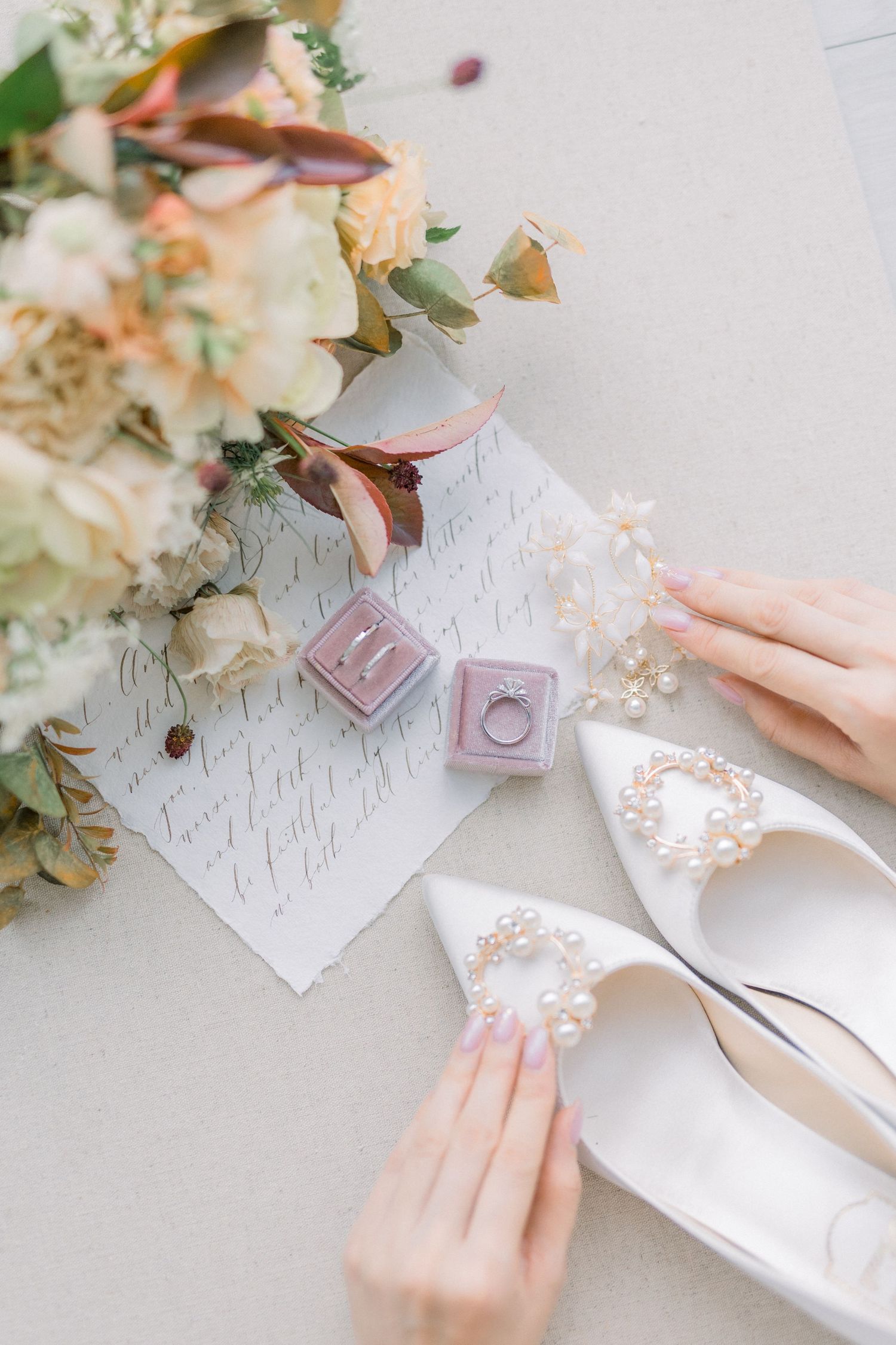 Elegant bridal details with wedding rings, shoes and floral arrangements on white surface.