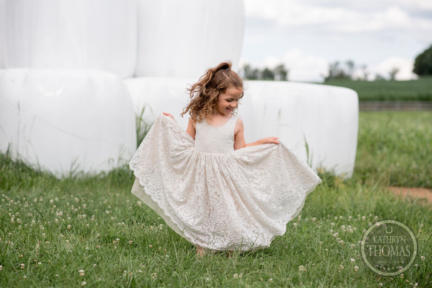 Young girl in white lace dress twirling among hay bales in field.