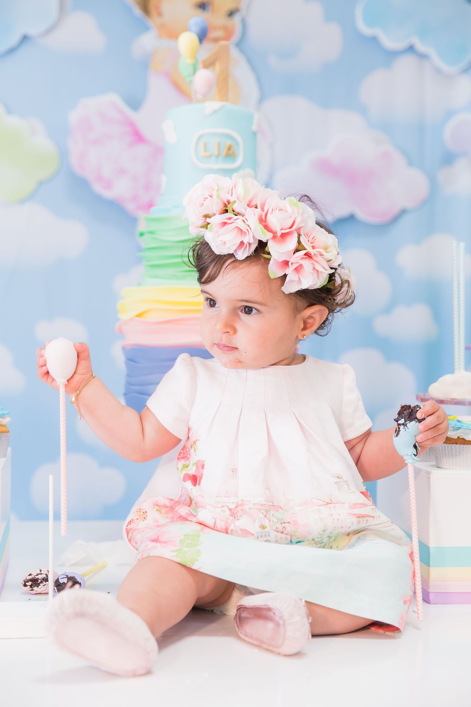 Baby in white dress with floral crown against pastel backdrop with rainbow cake.