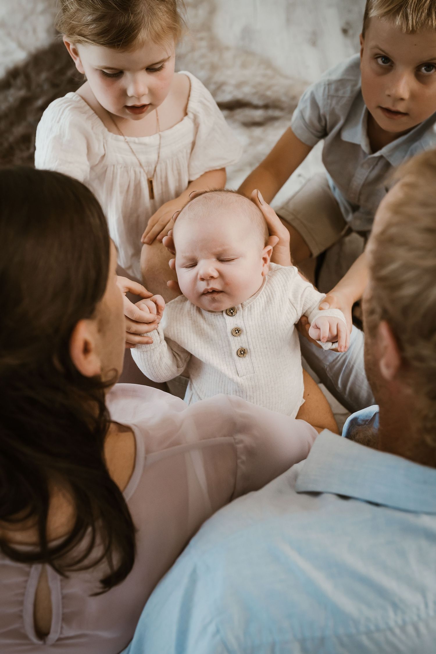 Intimate family moment as siblings gather around a small baby wearing a white henley onesie.
