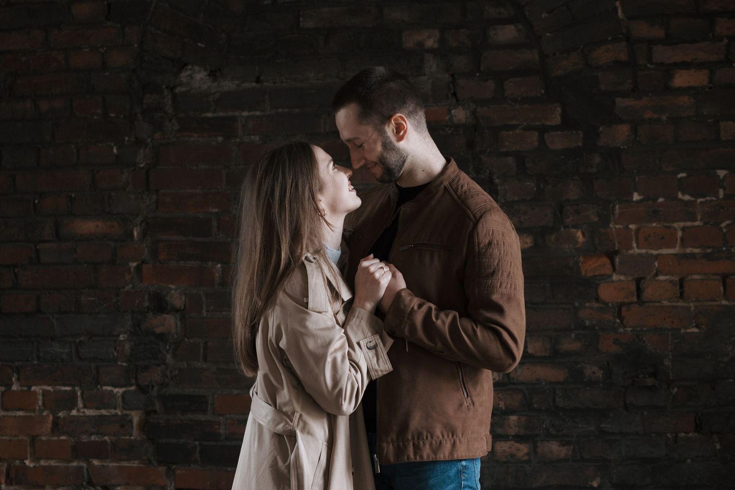Romantic couple embraces against rustic brick wall in casual winter attire.