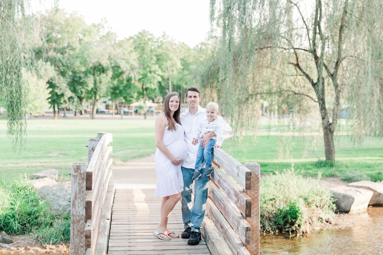 Family maternity portrait on rustic wooden bridge surrounded by weeping willows.