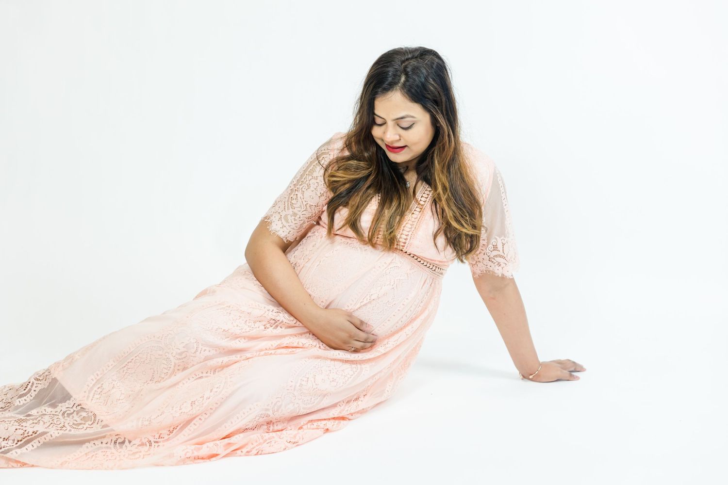 Expectant woman in blush pink lace maternity gown poses on white background.