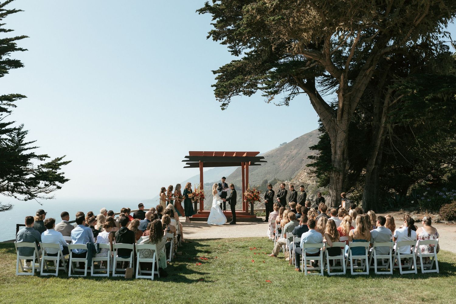 An outdoor coastal wedding ceremony setup with rows of white chairs and guests overlooking the ocean on a sunny day.