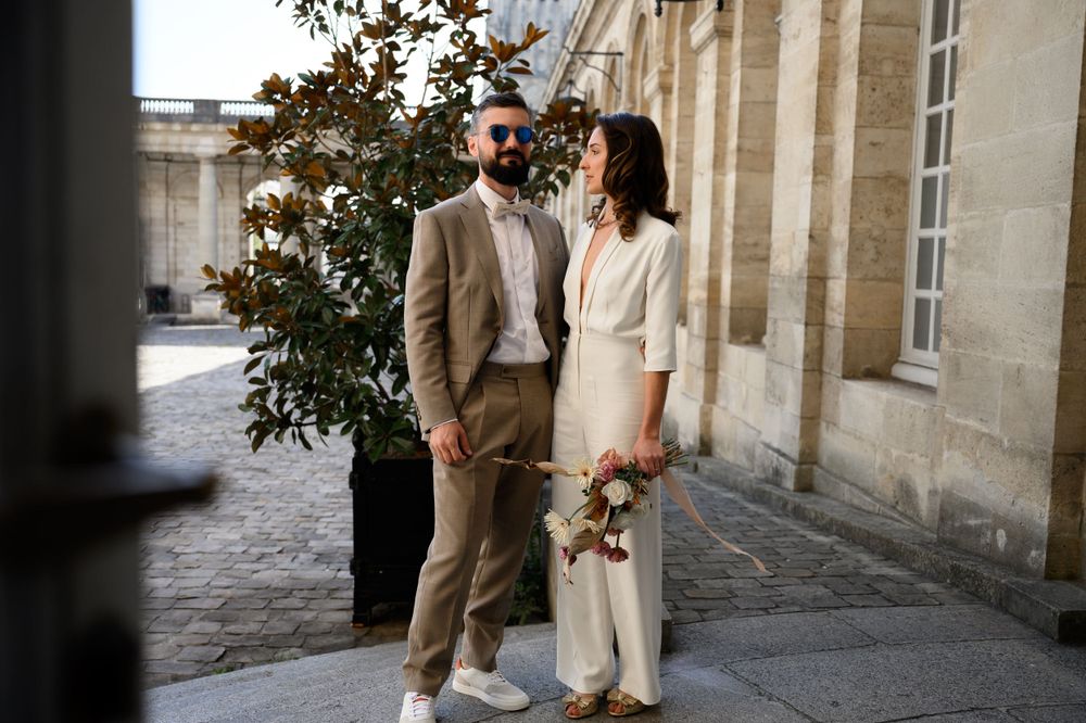 A couple in elegant attire stands together against a classic stone building exterior.