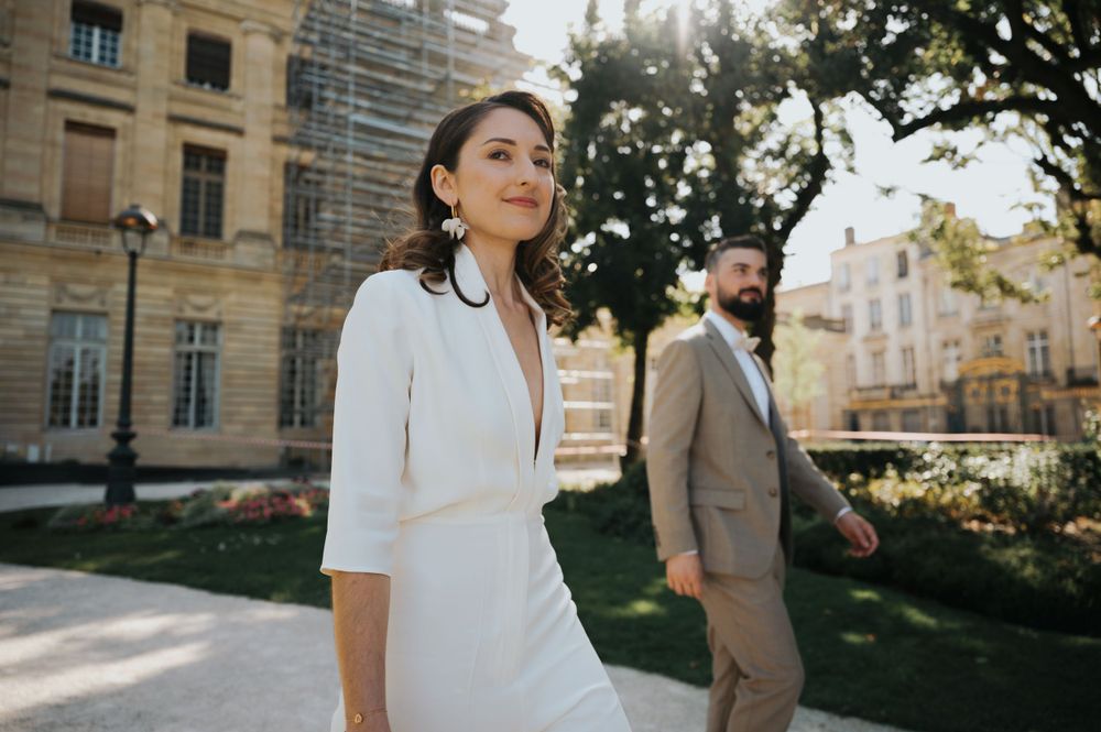 A couple in wedding attire walks outside a historic building on a sunny day.