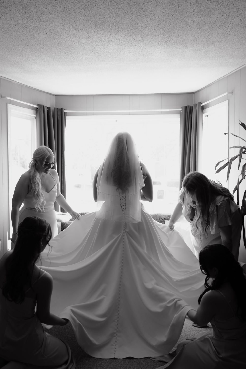 Black and white photo of a bride getting ready with her dress spread out against window light.