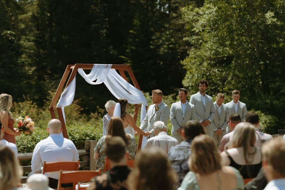 Outdoor wedding ceremony under wooden arch with white fabric decor and forest backdrop.