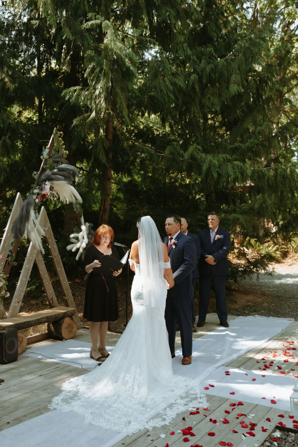 A wedding ceremony takes place outdoors under evergreen trees with rose petals scattered on a white aisle runner.