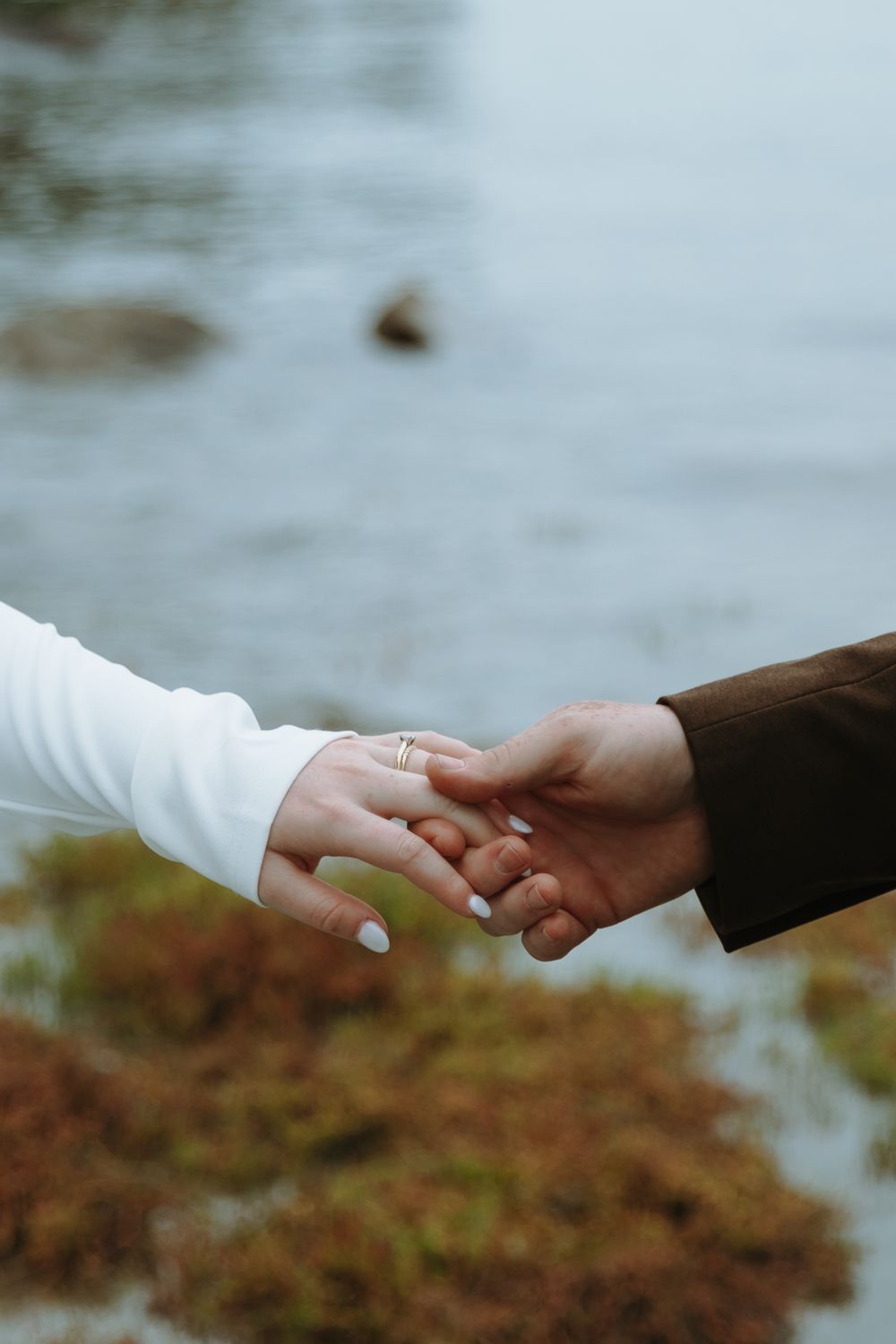 Two hands join together showing a wedding ring against a blurred waterfront background.