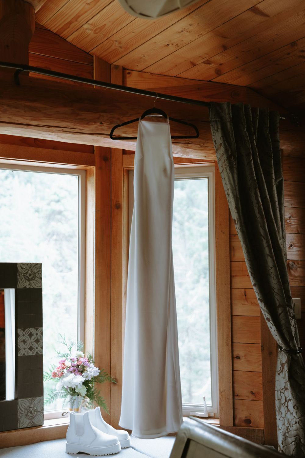 White wedding dress hanging in a rustic wooden cabin room with natural light streaming through windows.