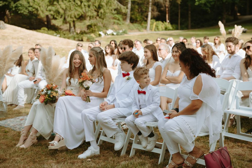 Wedding guests dressed in white seated on folding chairs at an outdoor ceremony.