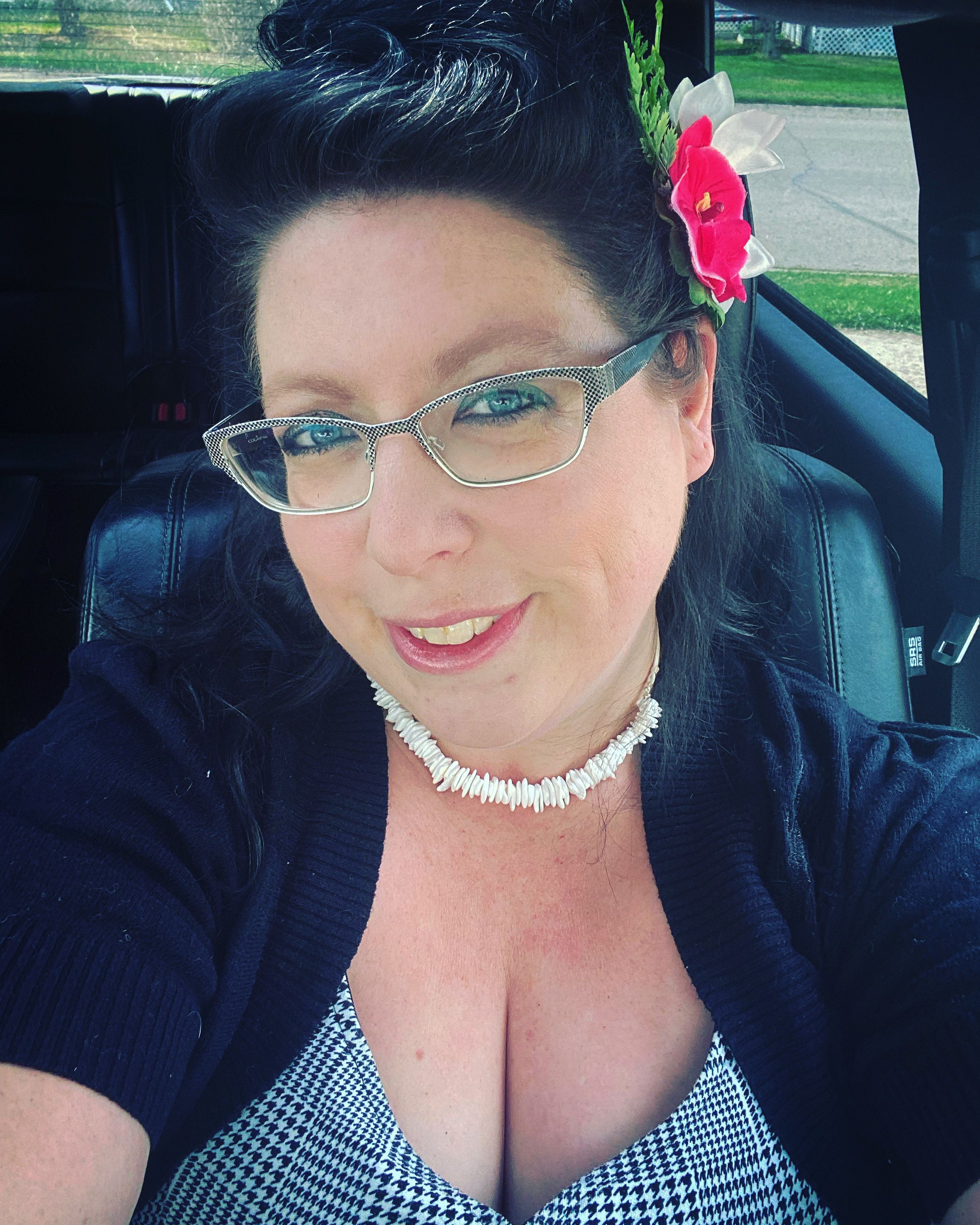 Woman in black cardigan with pink flower takes selfie in car.