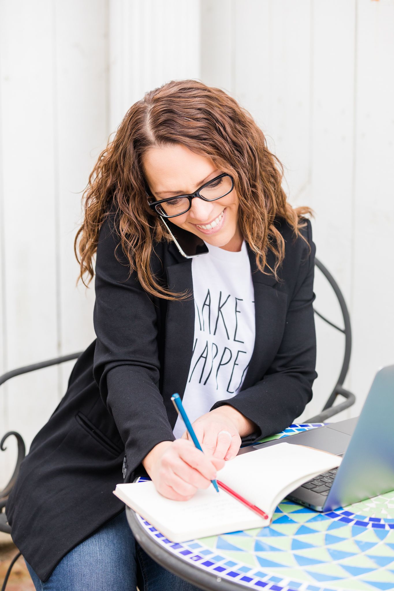 White woman with brown hair and glasses wearing white tshirt and black blazer talks on cell phone and writes in notebook