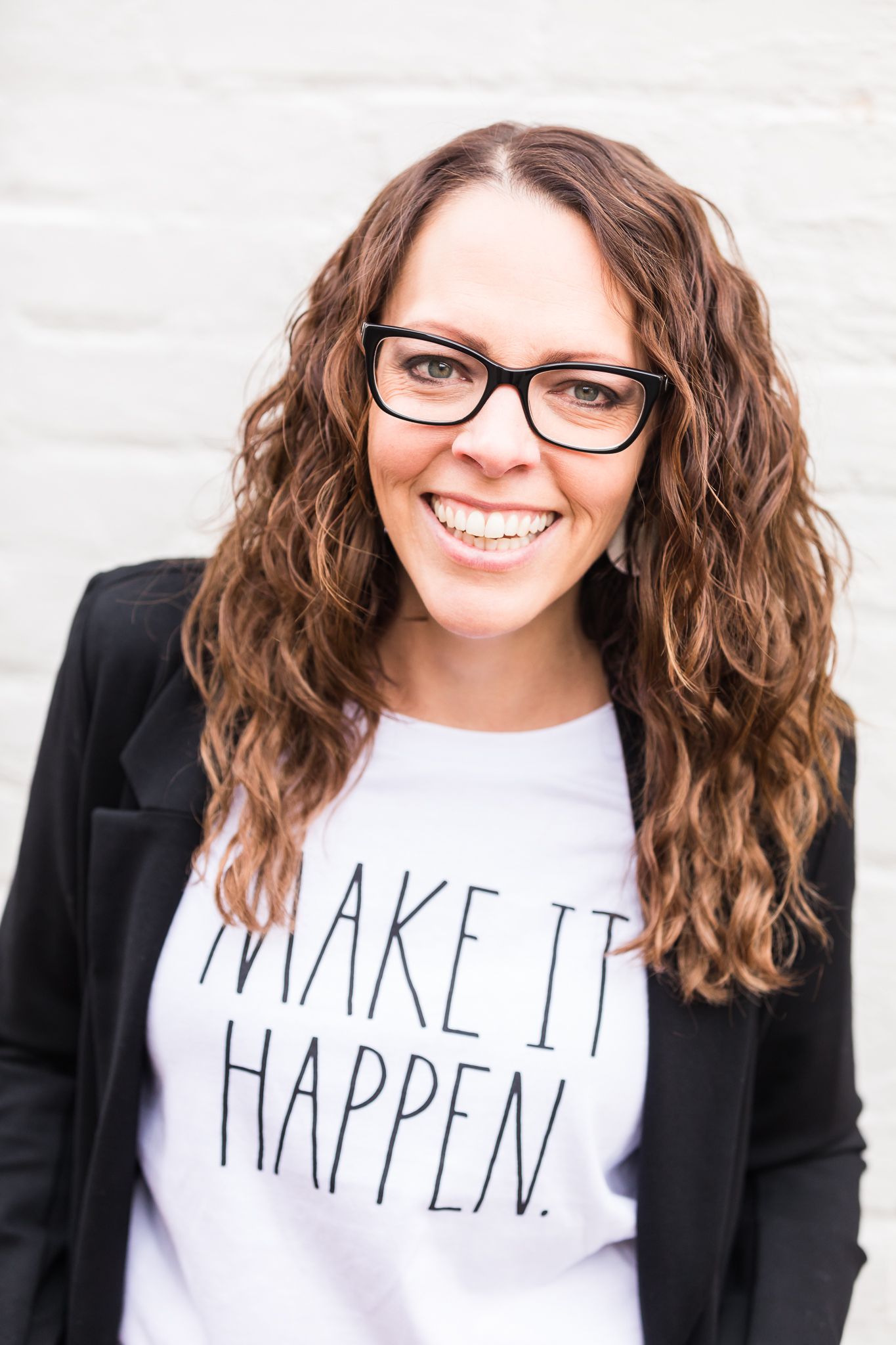 White female with brown hair and glasses poses against white wall wearing a white tshirt and black blazer