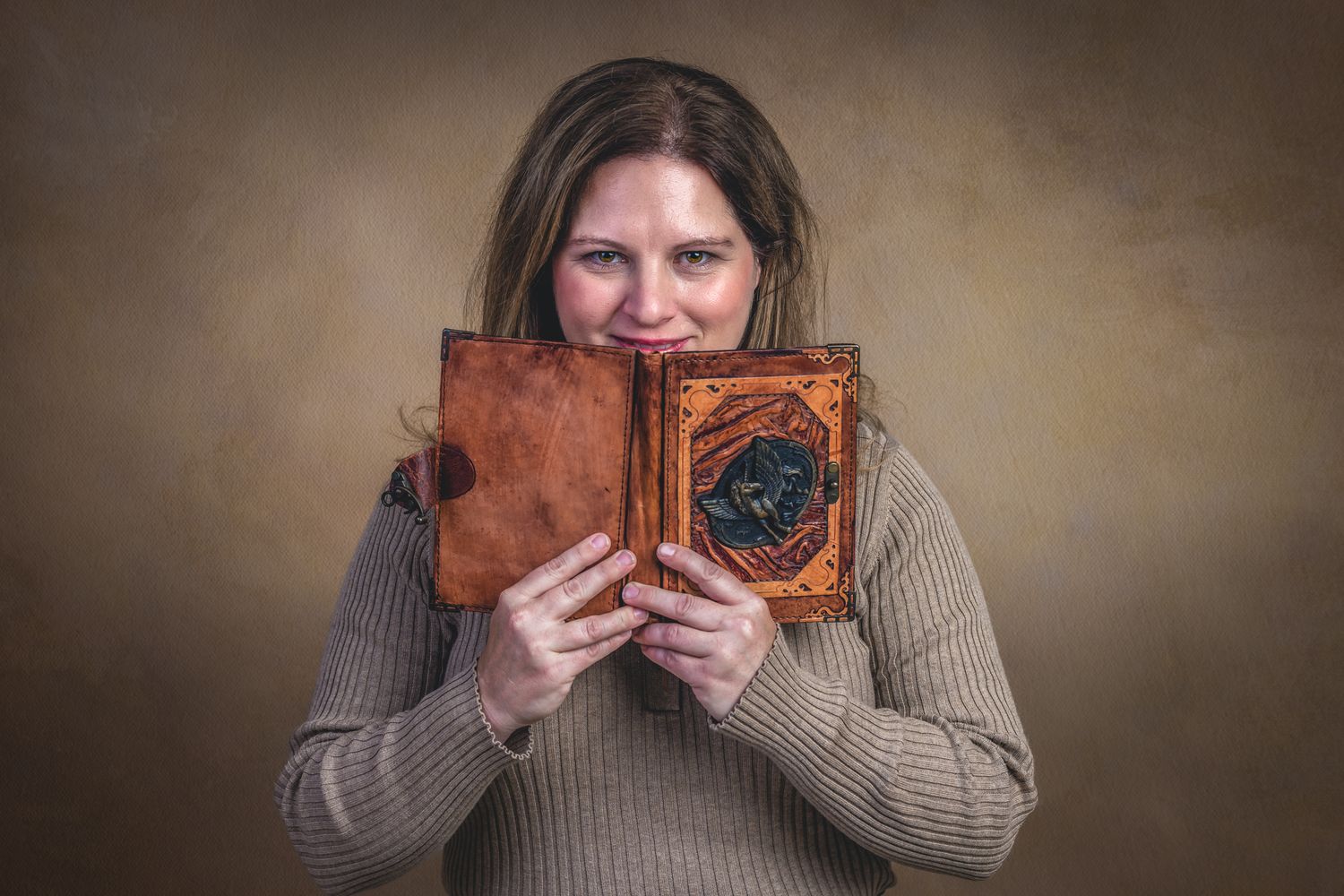 Woman smiling while holding an ornate vintage leather-bound book up to her face against a brown background.