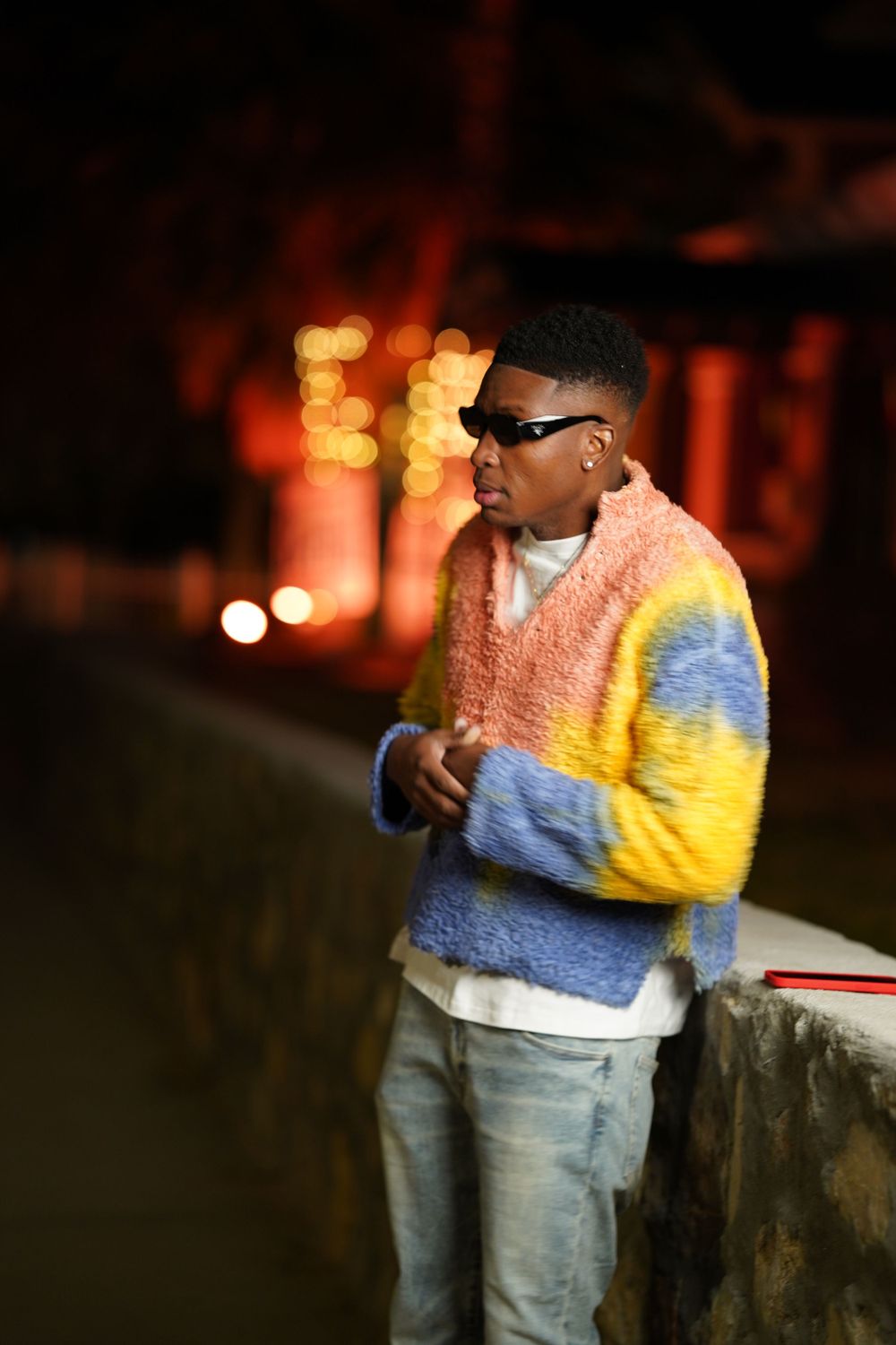 A person wearing sunglasses and a colorful pastel sweater poses against a dark background with bokeh lights.