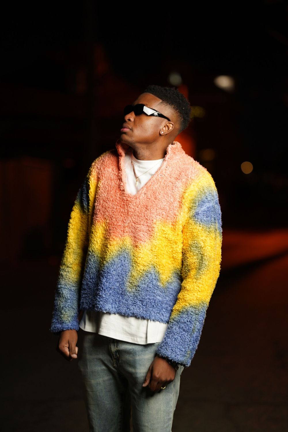 Figure wearing sunglasses and a colorblock sweater posed in moody nighttime lighting.