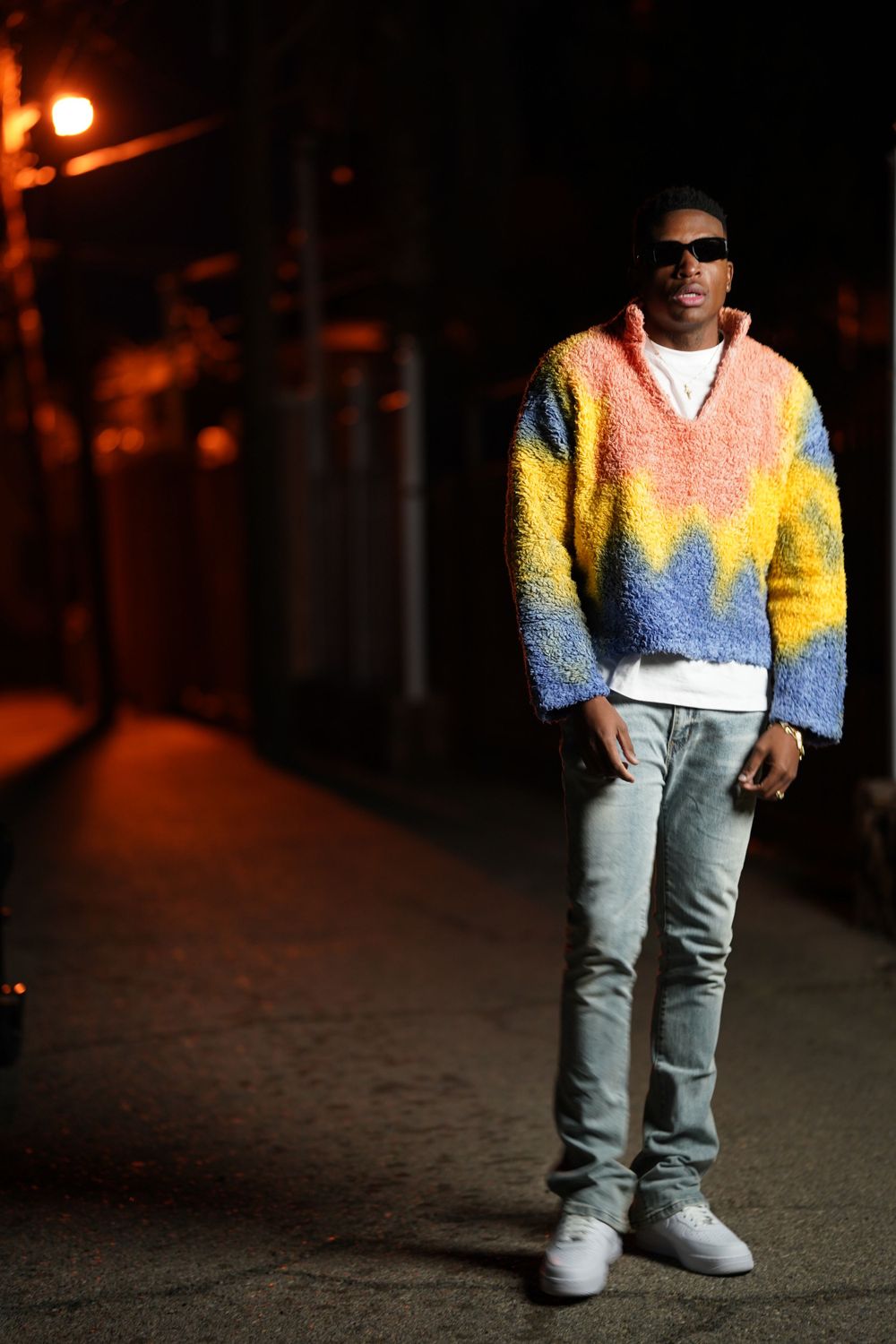 Person in a bright patterned sweater and light jeans posing under streetlights.
