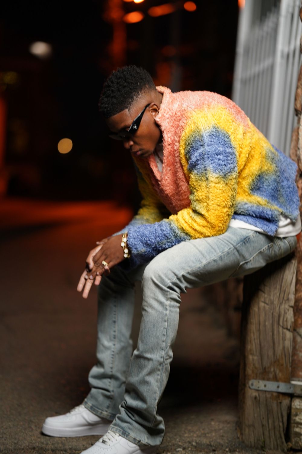 Figure in a multicolored fuzzy sweater and jeans leaning against a post at night.