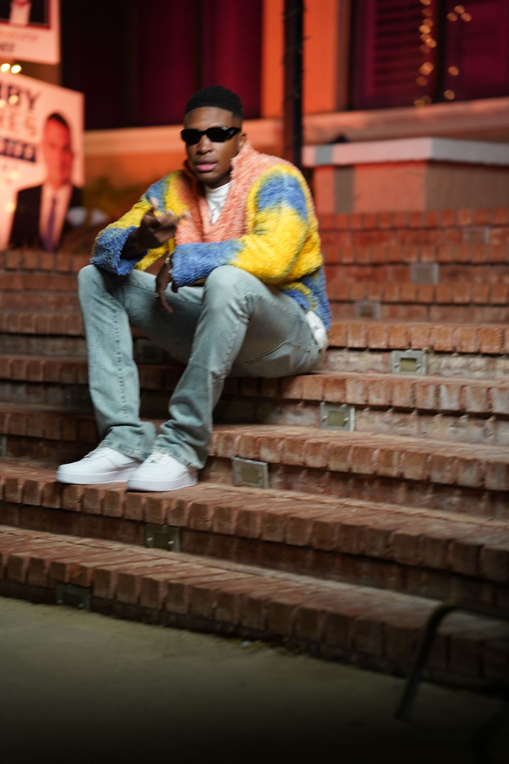 Someone sits on brick steps at night wearing a pastel multicolored sweater and light jeans.