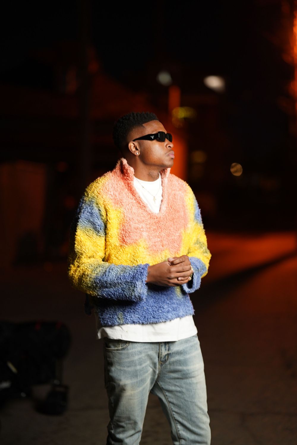 Individual wearing sunglasses and a colorful sweater standing in dramatic nighttime lighting.