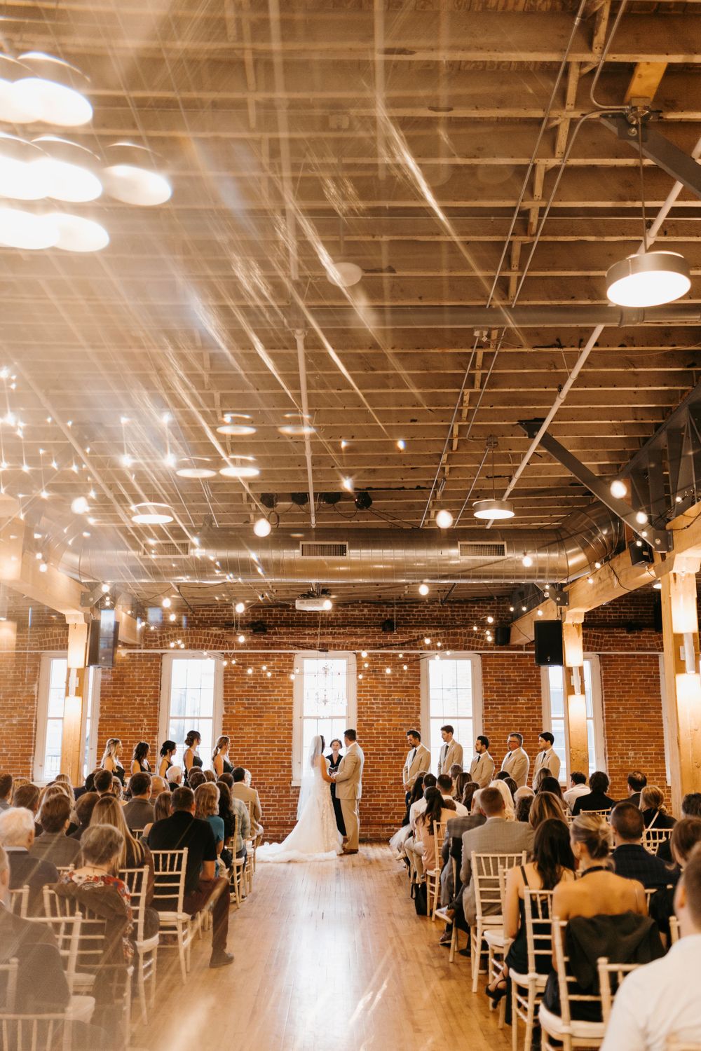 Sunlit wedding ceremony with couple at altar in brick-walled venue with string lights.