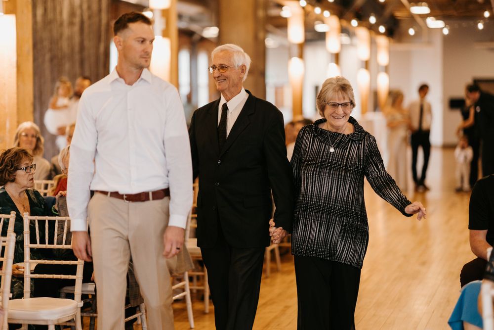 A young man escorts an older couple down the wedding ceremony aisle in an elegant venue.