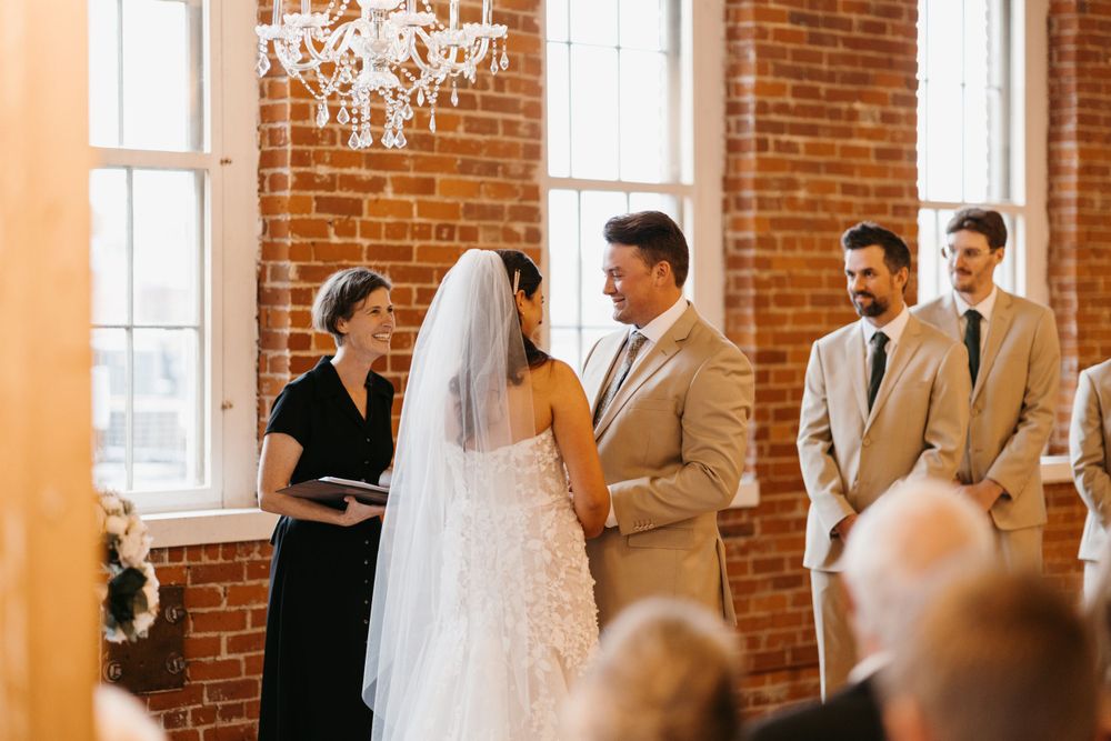 Bride and groom exchange vows before officiant in elegant brick-walled venue with chandelier.