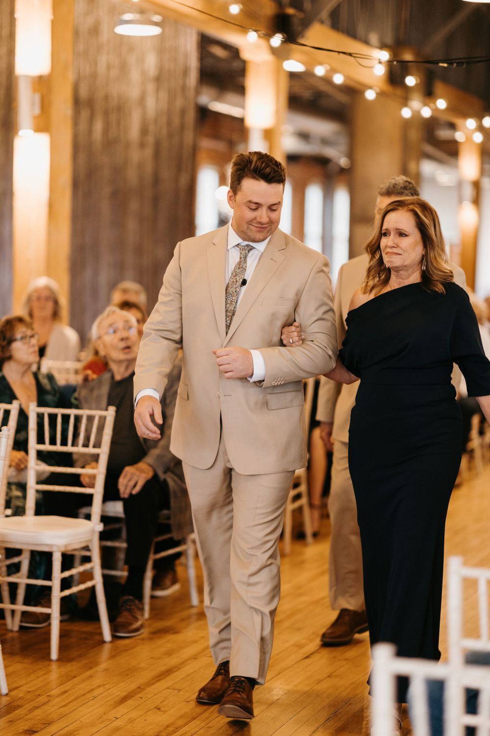 A groom in a tan suit walks his mother down the aisle at an indoor wedding ceremony.