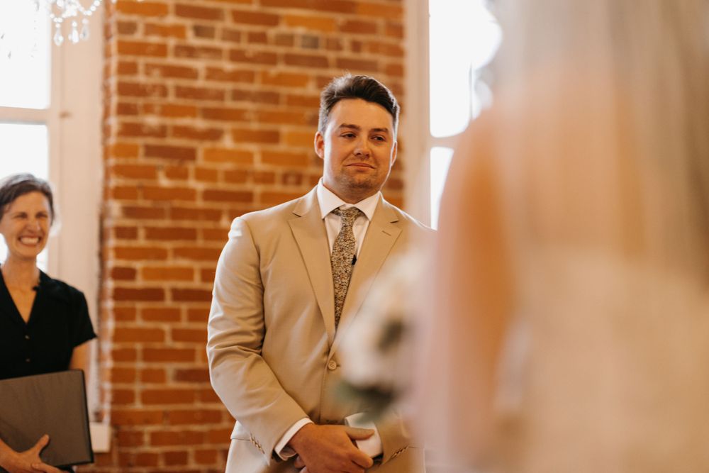 Groom in tan suit smiles warmly as bride approaches altar in brick-walled ceremony space.