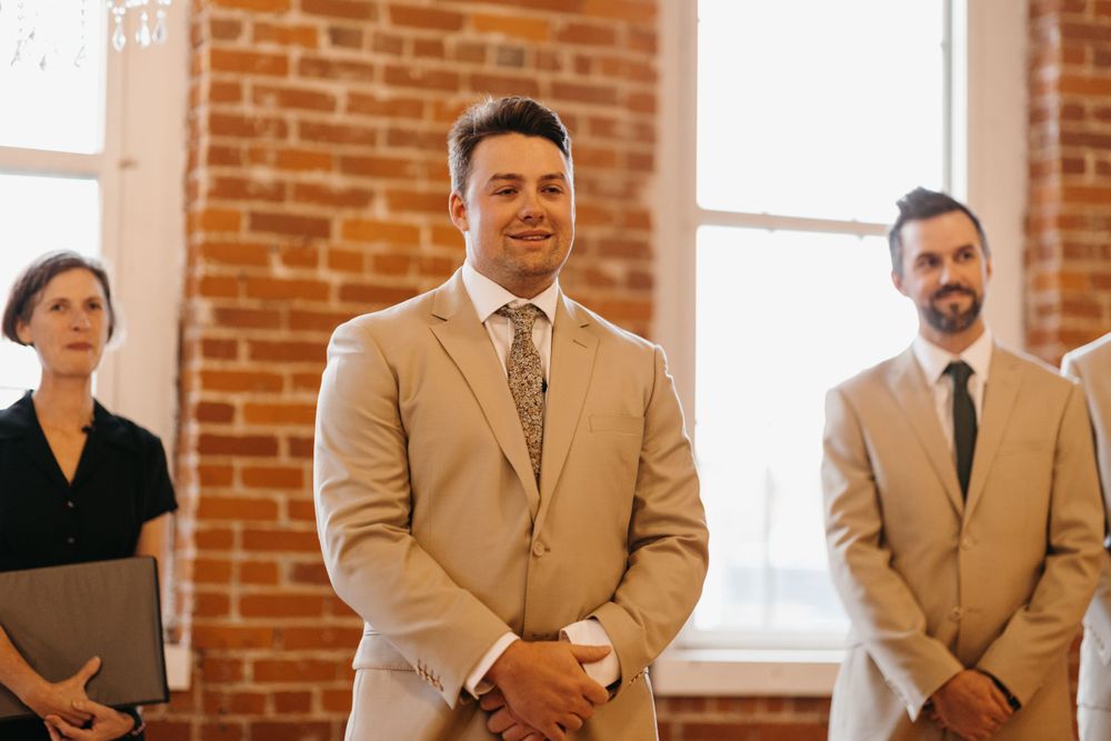 Groom in beige suit smiles at altar during wedding ceremony in brick-walled venue.