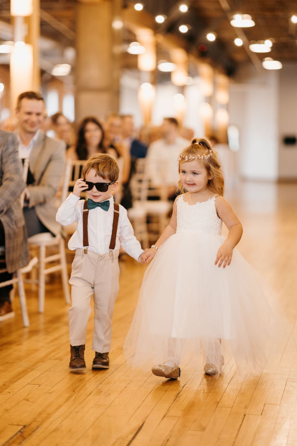 A ring bearer in suspenders and sunglasses holds hands with a flower girl in a white tulle dress.