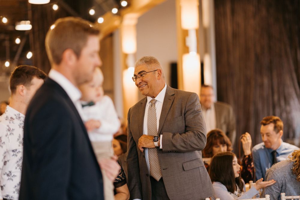 A man in a gray suit smiles warmly during a wedding reception dinner in a rustic venue.