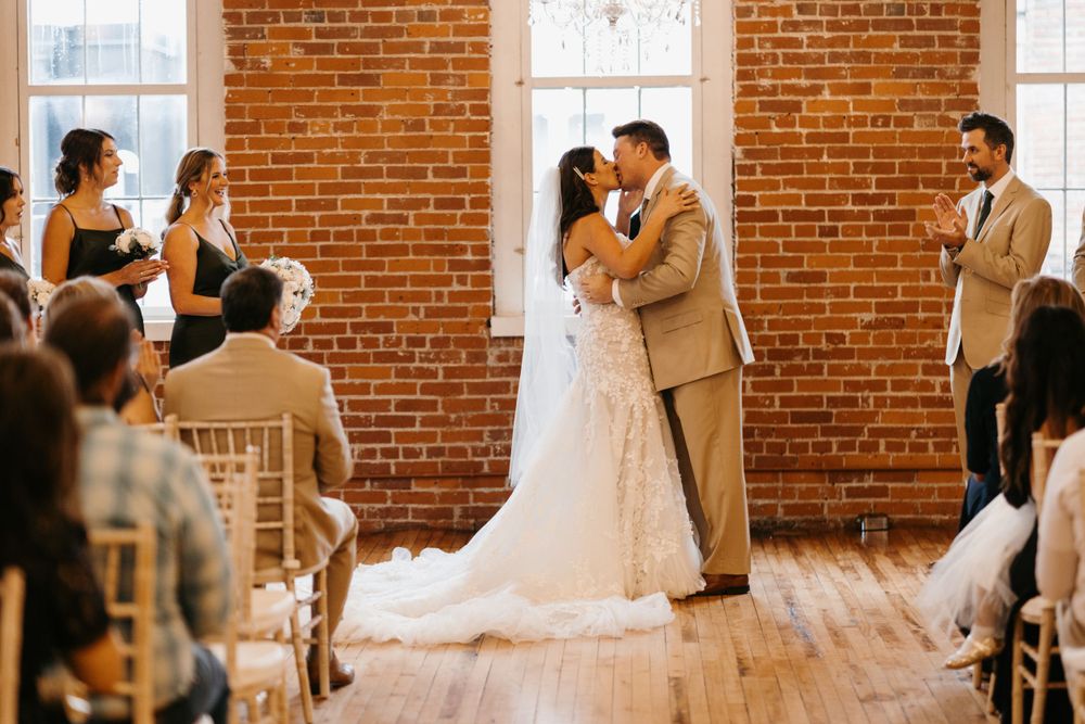 Bride and groom share first kiss at wedding ceremony in charming brick-walled event space.
