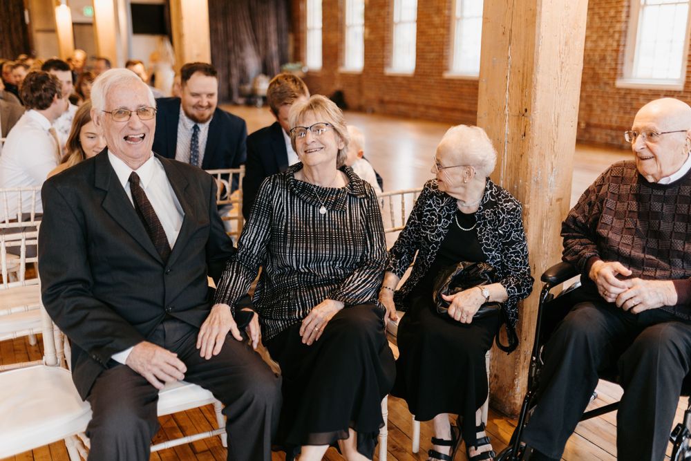 Smiling elderly wedding guests seated in white chairs during indoor ceremony celebration.