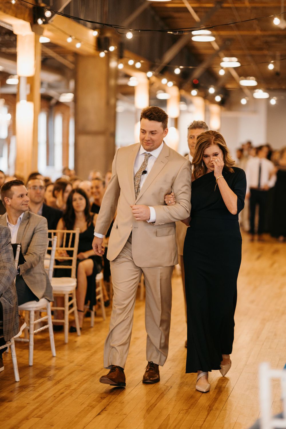 An emotional mother in a black dress is escorted down the aisle by her son at a wedding.