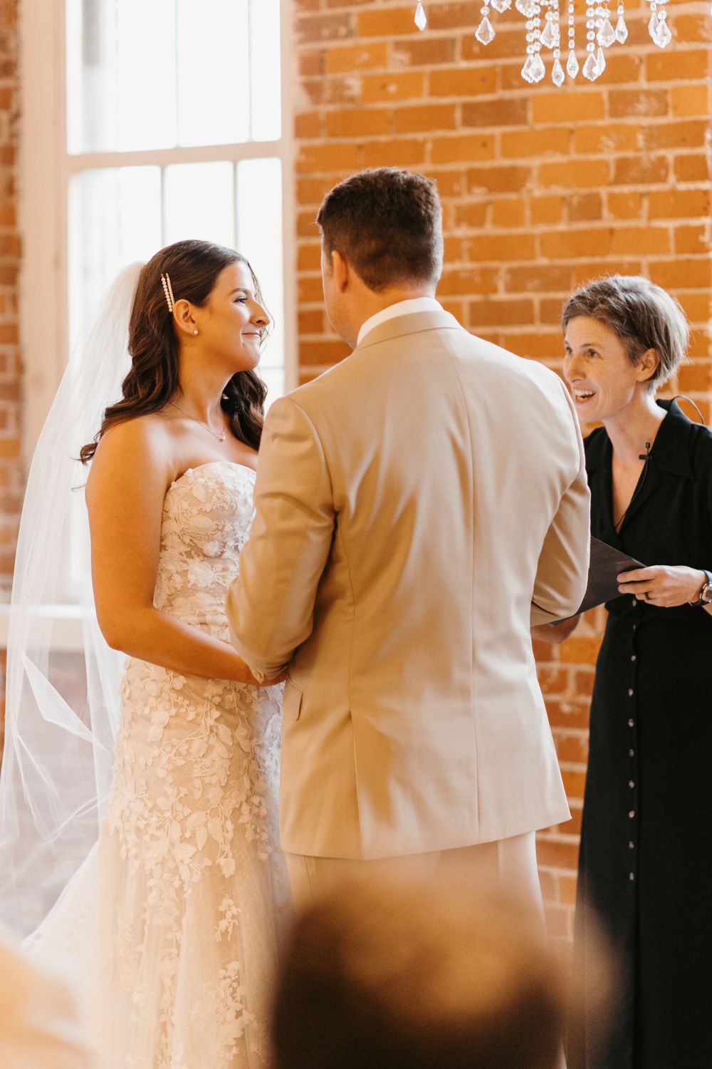 Bride in lace gown smiles at groom during wedding ceremony officiated near brick wall.