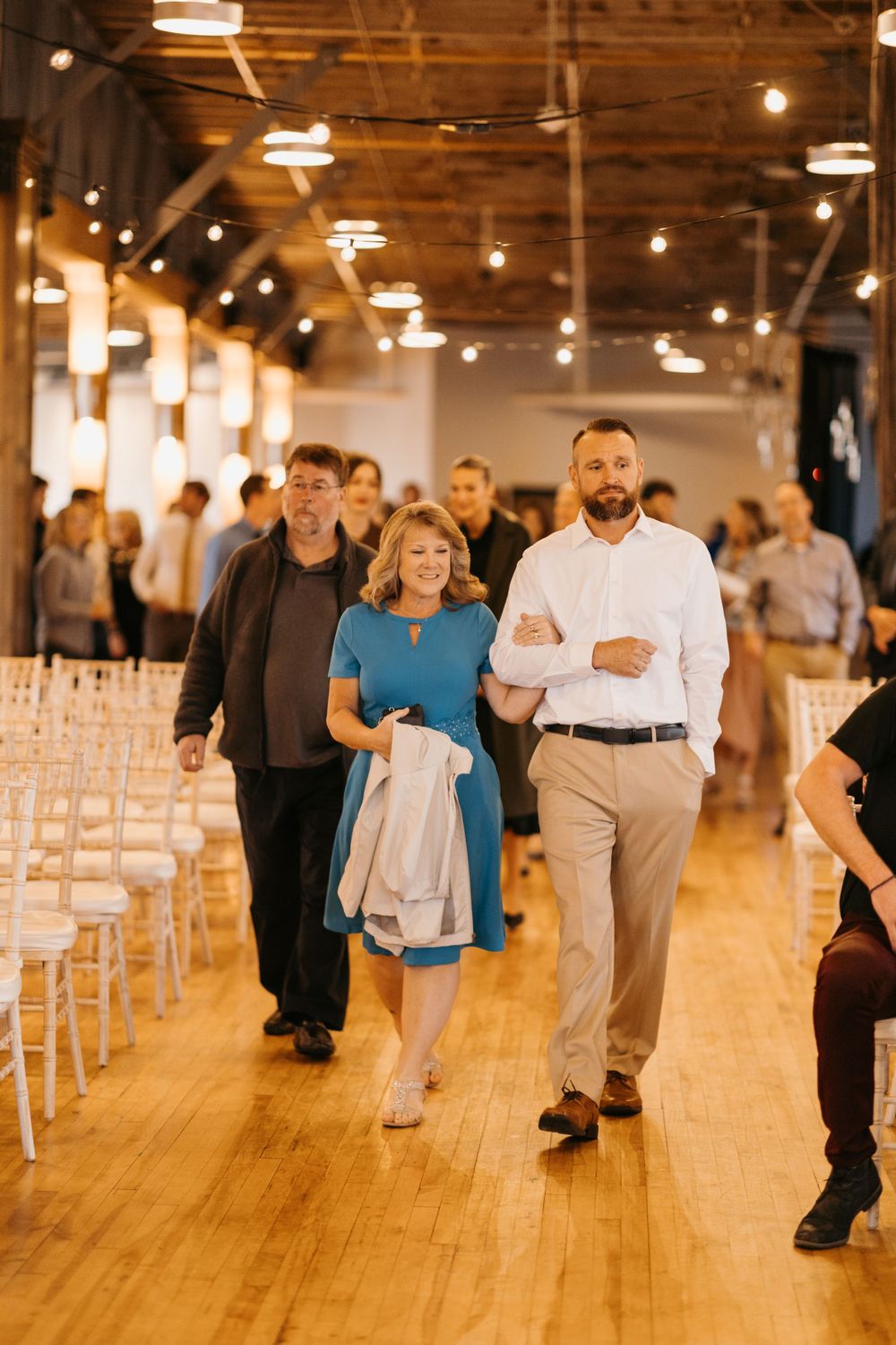 Groomsman escorts woman in teal dress down wedding ceremony aisle in warmly lit venue.