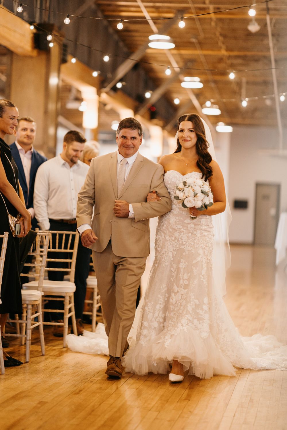 Bride in lace gown walks down aisle with father in tan suit holding white bouquet.