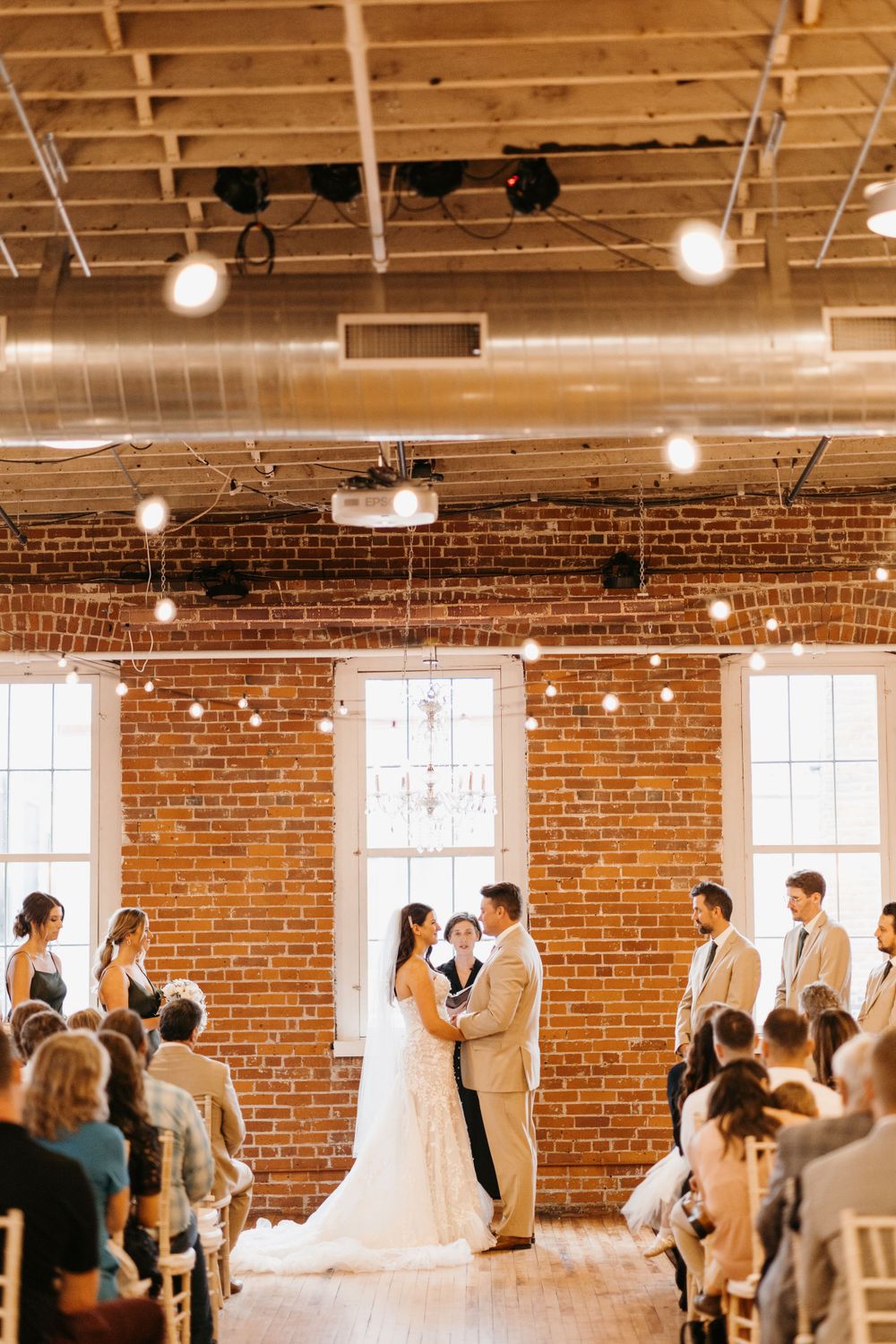 Wide shot of wedding ceremony in rustic brick venue with string lights and seated guests.