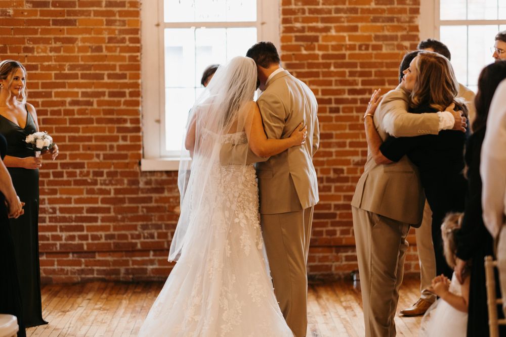 Bride and groom embrace at altar while guests hug nearby during wedding ceremony.