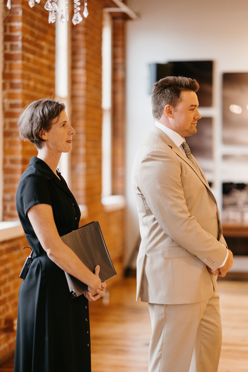 A wedding officiant in black holds a binder beside a groom in a tan suit at the altar.