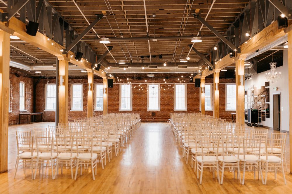 Empty wedding ceremony venue with white Chiavari chairs, exposed brick walls, and string lights.
