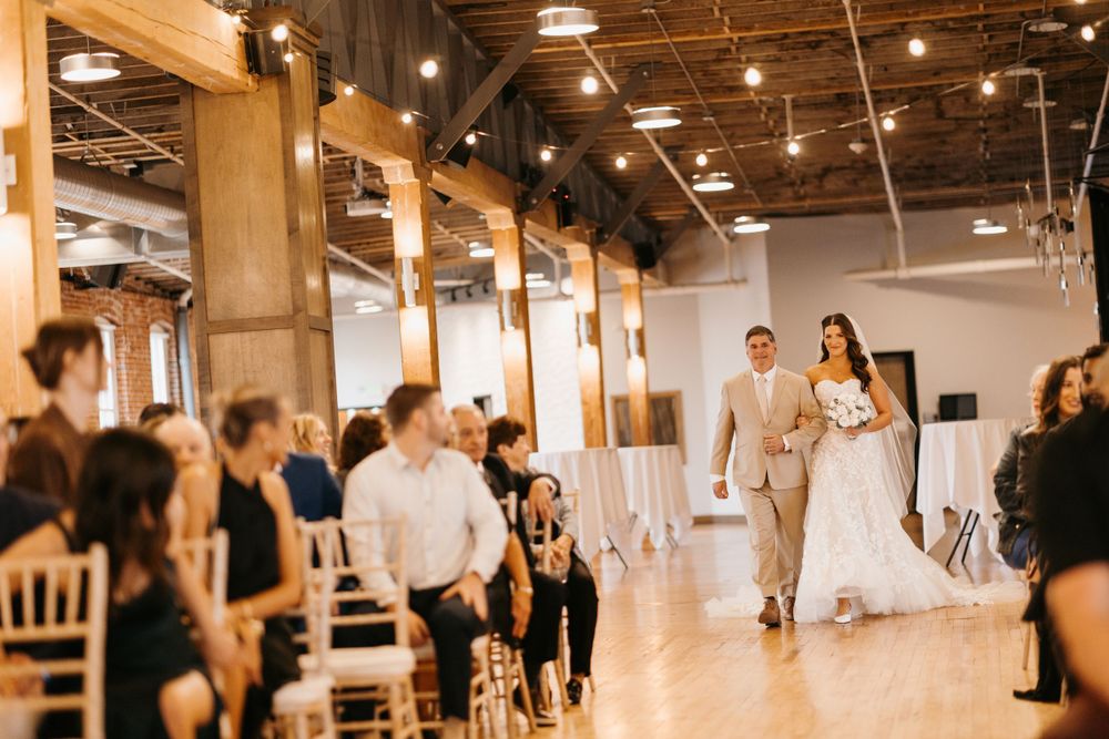 Bride walks down the aisle with escort in elegant indoor wedding venue with string lights.
