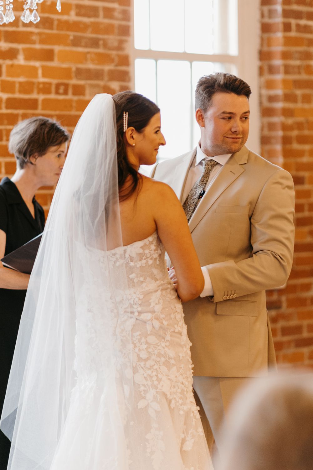 Groom in tan suit gazes at bride in lace gown during indoor wedding ceremony.