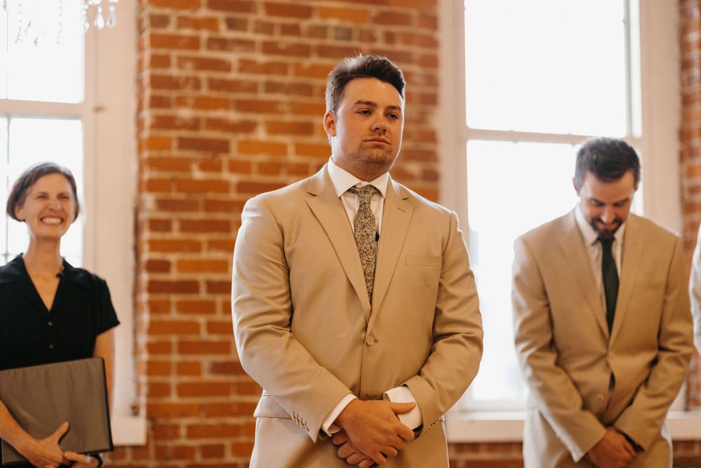 Groom in tan suit stands at altar with eyes closed during wedding ceremony near brick wall.